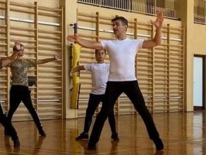 Na zdjęciu Jacek Guzowski, instruktor tańca towarzyskiego. Szczupły brunet w białym podkoszulku i czarnych spodniach, w pozie tanecznej tłumaczy kroki uczniom. Zdjęcie wykonane podczas zajęć.