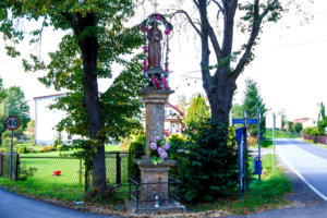 Przydrożna kapliczka w Krzęcinie ze stojącą na postumencie figurą Chrystusa Cierniem Ukoronowanego. W tle krzewy i drzewa.