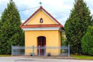Kapliczka Matki Boskiej Częstochowskiej w Facimiechu. Kapliczka to mały budynek z pomarańczową elewacją i czerwonym dachem, ogrodzony niskim płotem. Po obu stronach rosną wysokie krzewy, prawdopodobnie thuje.
