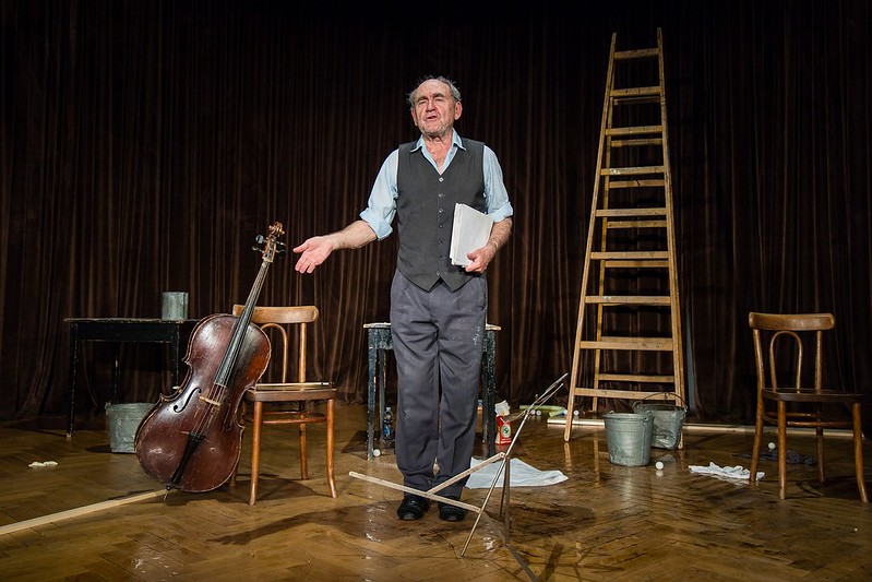 Na scenie teatralnej Jan Peszek, niski, szczupły, starszy mężczyzna, garniturowe spodnie, kamizelka, niebieska koszula. Na scenie stoją drewniane: drabina, krzesła, stoły oraz wiolonczela.