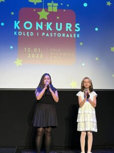 Dwie dziewczynki około dziesięcioletnie stoją na scenie i śpiewają.