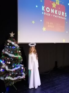 Dziewczynka 9 lat w stroju aniołka stoi na scenie przy udekorowanej choince.