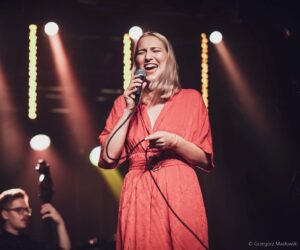 Instruktorka wokalu, wokalistka. Młoda dziewczyna w czerwonej sukience, blond włosy, śpiewa na scenie do mikrofonu. W tle reflektory, ciemne tło.