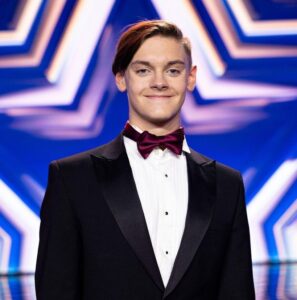 Uśmiechnięty młody chłopak w czarnym smokingu, białej koszuli i bordowej muszce. Tło niebiesko-białe.