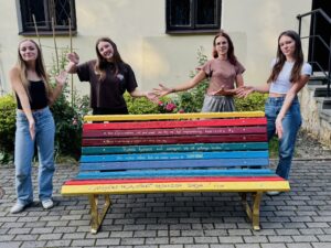 Na zdjęciu cztery młode kobiety stoją wokół kolorowej ławki pomalowanej w poziome pasy w różnych barwach. Na deseczkach widnieją inspirujące cytaty. Wszystkie uśmiechają się i gestem rąk prezentują swoją pracę.