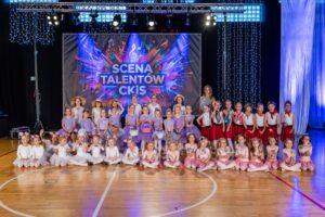 Na zdjęciu duża grupa baletniczek, dziewczynek od 5 lat po nastolatki. Dziewczynki pozują w strojach baletowych. W pierwszej linii, młodsze dziewczynki siedzą, a za nimi, w drugiej linii dziewczynki stoją. Wśród nich stoi instruktorka. W tle widnieje baner z napisem "Scena talentów CKiS".