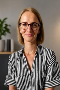 Zdjęcie przedstawia kobietę, od pasa w górę. Kobieta ubrana jest w bluzkę w paski, ma czarne okulary, włosy blond do ramion. Delikatnie się uśmiecha.