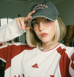 Zdjęcie przedstawia kobietę, ujętą od pasa w górę, w sportowej biało-czerwonej bluzie i szarej czapeczce z daszkiem. Kobieta ma okulary i blond włosy do ramion. Prawą ręką, uniesioną do góry trzyma daszek swojej czapki.