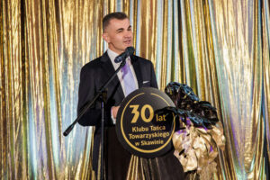 Mężczyzna, w eleganckim, czarnym garniturze, stoi za mównicą z napisem "30 lat Klubu Tańca Towarzyskiego w Skawinie". W tle postaci wisi złota tkanina.