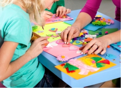 Na zdjęciu fragment stołu z niebieskim blatem, na którym rozłożone są różnokolorowe materiały plastyczne i wyklejanki. Dziecięce dłonie przyklejają elementy na różowej kartce.